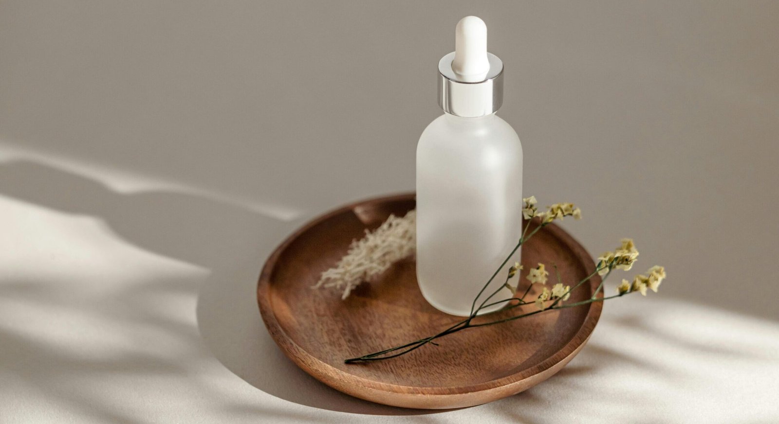 Elegant skincare bottle displayed on a wooden saucer with dried flowers and soft shadows.